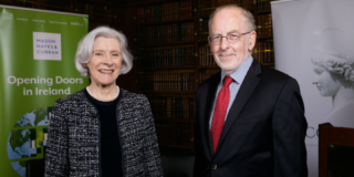 A woman (left) with grey hair in a bob and a man with short grey hair (left), wearing glasses, face the camera smiling. She wears a grey formal jacket and he wears a black suit with blue shirt and red tie.
