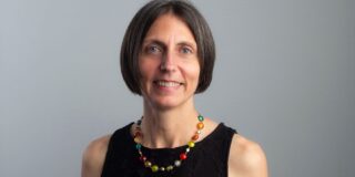 A headshot of a woman with dark brunette hair in a bob, standing against a grey background. She wears a black sleeveless dress and a coloured beaded necklace.