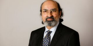 A headshot of a man with short greying hair and a beard, wearing a dark coloured suit, navy patterned tie and white shirt, against a grey background
