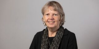 A headshot of a woman with short grey hair, wearing a black dress, dangling earrings and a dark patterned scarf, against a grey background.