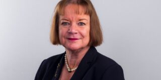 Headshot of a woman against a grey backdrop, she has short auburn hair and a fringe, she wears a black blazer, pearl necklace
