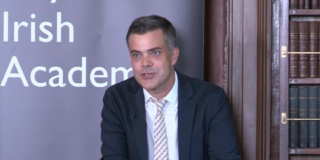 A man with dark hair, wearing a navy suit with white shirt and beige striped tie, is looking off camera as he addresses an audience. He stands in front of a Royal Irish Academy banner to his left and a bookshop with leather bound books on his right.