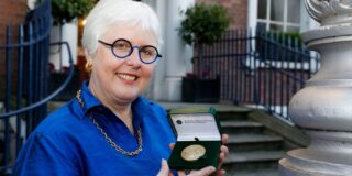 A woman with short white cropped hair, wearing black and blue rimmed glasses with a circular shaped lens, a blue silk blouse and gold chain necklace looks toward the camera smiling. She is holding a presentation box with a gold medal inside, in the background are granite steps with railings on either side, leading up to a redbrick building.