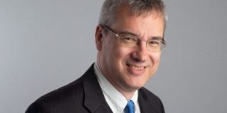 A headshot of a man with short grey hair, wearing glasses and a dark coloured suit with a white shirt and blue tie. He stands against a grey background.